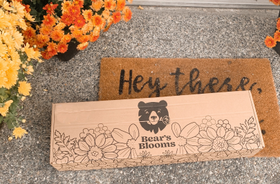 A long cardboard box with the Bear’s Blooms logo sits on a front doorstep. It is surrounded by potted chrysanthemums in orange and yellow.